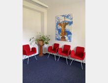 Ein heller Warteraum mit vier roten Stühlen, einem Topfpflanzenständer in einer Ecke und einem großen Wandbild der goldenen Siegessäule vor blauem Himmel. Der Boden ist mit dunkelblauem Teppich ausgelegt und die Wände sind weiß.