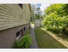 Schmaler Seitenbereich neben einem Wohnhaus mit hell verklinkerter Fassade und kleinem Kellerfenster, entlang eines gepflasterten Weges. Rechts grenzt eine grasbewachsene Fläche mit dichtem Strauch- und Baumbewuchs an, im Hintergrund steht ein graues Metalltor oder Schuppen vor einem Waldsaum.