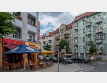 Straßenszene in einer urbanen Umgebung mit mehrstöckigen Wohnhäusern im Altbaustil, Restaurants und Cafés im Erdgeschoss. Vor einem mexikanischen Restaurant mit roten Wänden und einem blauen Sonnenschirm stehen Tische und Stühle, daneben geparkte Autos und Fahrräder; eine Person fährt mit dem Fahrrad auf der Straße.