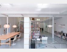 Modernes, helles Büro mit minimalistischer Einrichtung, einschließlich eines großen Holztischs mit weißen Stühlen, einem Glas-Schreibtisch, Bücherregalen voller Bücher und einer offenen Treppe zu einer oberen Ebene. Die weißen Wände und der graue Boden schaffen eine klare, strukturierte Atmosphäre.