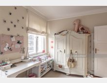 Helles Kinder- oder Jugendzimmer mit einem großen weißen Kleiderschrank, einem Schreibtisch mit Schreibtischlampe und einer Pinnwand an der Wand. Ein Fenster mit beigefarbener Jalousie lässt Tageslicht herein, und der Raum ist mit persönlichen Gegenständen dekoriert.