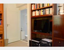 Ein gemütlicher Innenraum mit einem hölzernen Bücherregal, das Bücher und einen Flachbildfernseher enthält. Im Vordergrund steht Bürotechnik, und im Hintergrund führt eine offene Tür zu einem hell beleuchteten Flur.