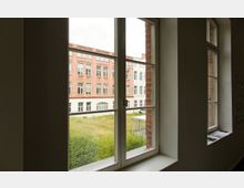 Blick aus einem Innenraum durch ein Fenster auf ein Backsteingebäude mit weißen und roten Fassadenelementen sowie zahlreichen Fenstern. Der Außenbereich zeigt eine Wiese mit vereinzelten Büschen und einem kleinen Überdachungsbau.