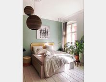 Helles Schlafzimmer mit einem Holzbett und hellem Bettzeug, grün gestrichener Wand, zwei kugelförmigen Deckenlampen und einem großen Fenster mit Vorhängen. Im Raum befinden sich eine Zimmerpflanze in einem Topf und ein moderner Kunstdruck an der Wand.