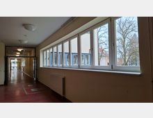 Langer Innenflur eines öffentlichen Gebäudes mit rotem Boden, Deckenleuchten und einer Reihe großer Fenster entlang der rechten Seite. Durch die Fenster sind ein gegenüberliegendes Gebäude sowie Bäume im Außenbereich zu sehen.