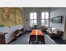 Ein Wohnzimmer mit altmodischem Dekor, einschließlich einer großen Wandkarte, einem Tisch mit dekorierter Tischdecke und einem Sofa an der rechten Seite. Zwei hohe Fenster lassen Tageslicht herein; im Hintergrund sind Möbelstücke und Dekorationsobjekte zu sehen.