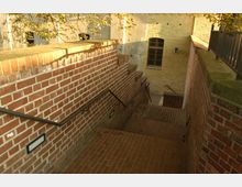 Eine Treppe aus roten Ziegelsteinen führt zwischen Ziegelwänden hinunter, mit schwarzen Geländern auf beiden Seiten. Im Hintergrund sind Wände und Fenster eines Gebäudes aus hellbeigem Ziegelwerk zu sehen, umgeben von gelbem Laub im Sonnenlicht.