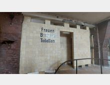 Innenansicht eines Gebäudes mit einem Bereich, der mit beigen Fliesen verkleidet ist und den Schriftzug "Frauen Brausen Toiletten" trägt. Rechts sind Rohre sichtbar, links ist eine Wand aus rotem Backstein.