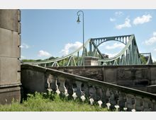 Grüne Stahlbrücke mit geschwungener Struktur und großen Bögen, im Vordergrund ein steinernes Geländer mit Verzierungen und daneben eine Laterne; im Hintergrund Bäume und blauer Himmel mit einigen Wolken.