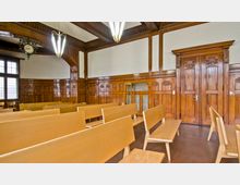 Ein Innenraum mit Holzwänden und einer getäfelten Decke, versehen mit schlichten Bänken aus hellem Holz. Ein großes Doppelfenster mit Buntglas und eine Wanduhr sind neben einer doppelflügeligen Holztür sichtbar.