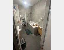 Modernes Badezimmer mit grauen Boden- und Wandfliesen, Badewanne mit Glas-Duschabtrennung und Handbrause sowie Handtuchheizkörper an der Stirnwand. Links stehen Waschmaschine und Waschbecken, rechts ein Holzschrank mit Wäschekorb; ein kleiner Teppich liegt vor der Wanne.