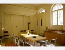 Heller Besprechungsraum mit großen Fenstern und hohen, cremefarbenen Türen. In der Mitte steht ein rechteckiger Holztisch mit mehreren Stühlen, darauf eine Vase mit gelben und orangefarbenen Tulpen; im Hintergrund sind Schränke und eine Garderobe.