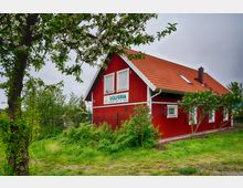 Ein rotes Holzgebäude mit weißen Fensterrahmen und einem roten Ziegeldach, umgeben von grüner Vegetation und Bäumen mit Blüten. Auf der Fassade befindet sich ein Schild mit der Aufschrift "Golferia", die Landschaft wirkt ländlich und naturbelassen.