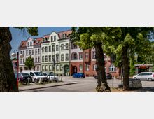 Straßenszene mit historischen Gebäuden, deren Fassaden in Pastelltönen und Ziegelstein gestaltet sind. Im Vordergrund stehen mehrere Bäume und geparkte Autos entlang der Straße.