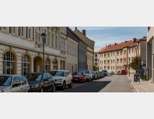 Straßenszene mit geparkten Autos auf beiden Seiten, flankiert von überwiegend historischen Wohngebäuden mit verzierten Fassaden und einfachen Mehrfamilienhäusern mit roten Ziegeldächern; Laternenmast und Gehwege erkennbar.