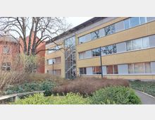 Mehrstöckiges Gebäude mit einer Fassade aus gelbem Klinker und großen Fenstern, das von einem kleinen Garten mit Büschen, Bäumen und einem Gehweg umgeben ist. Links ist ein rotes Backsteingebäude sichtbar, und in der Mitte des Bildes führt ein Eingang mit Glasfront in das Gebäude.