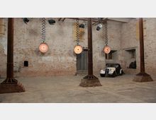 Industriehalle mit unverputzten Backsteinwänden, vier metallenen Säulen und hängenden Heizstrahlern. Im Hintergrund steht ein weiß-schwarzer Oldtimer neben einer Holztür, umgeben von weiteren kleinen Fenstern und groben Wandelementen.