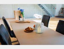 Heller, moderner Essbereich mit großem rechteckigem Tisch und dunklen Polsterstühlen, dekoriert mit Kerzen, Blumen und Frühstücksgebäck. Im Hintergrund sind ein offenes Treppengeländer, helle Holzböden und ein einzelner Stuhl an der Wand zu sehen.