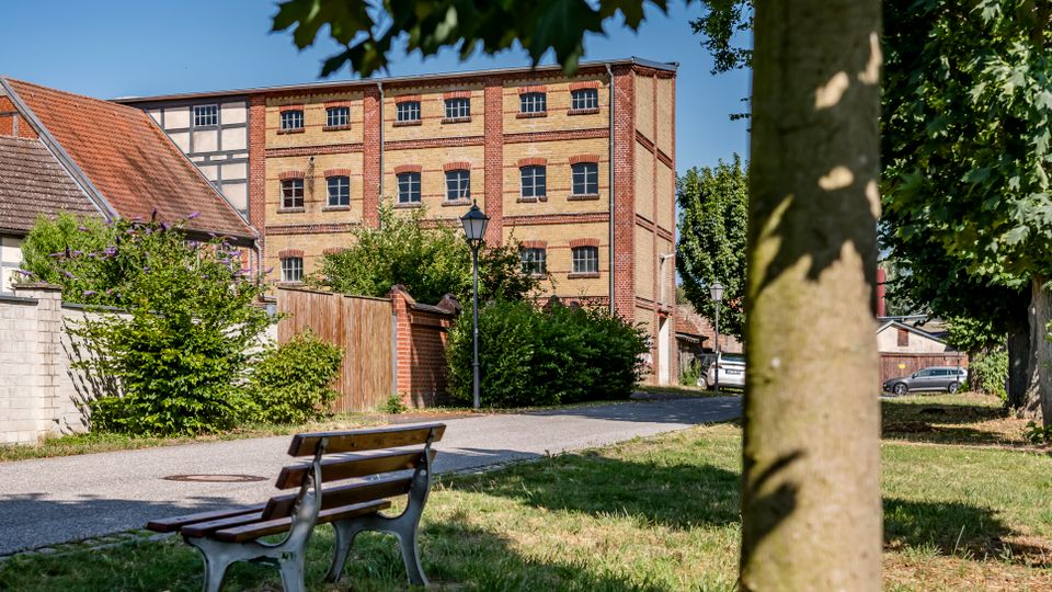 Blick auf einen kleinen Grünstreifen mit Parkbank und Bäumen neben einem Fußweg, der zu einem mehrstöckigen Backstein-Industriegebäude mit vielen Fenstern führt. Links stehen Fachwerk- und Ziegelhäuser, entlang des Weges sind Büsche und eine Straßenlaterne zu sehen.