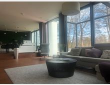 Wohnzimmer mit großen Panoramafenstern, die auf eine baumreiche Außenlandschaft blicken. Der Raum ist modern eingerichtet mit einem grauen Sofa, einem schwarzen Couchtisch, einem grauen Sessel und einer weißen Theke im Hintergrund; Holzboden und ein heller Teppich vervollständigen das Ambiente.