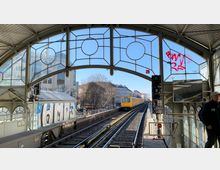 Bahnhof mit überdachter Bahnsteighalle und großen Glasfenstern mit geometrischen Mustern, durch die Stadthäuser und Bäume bei klarem Himmel sichtbar sind; ein gelber U-Bahn-Wagen fährt in den Bahnhof ein.