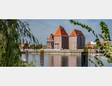 Ziegelbauten mit roten Satteldächern stehen am Ufer eines Hafenbeckens, umgeben von modernen und historischen Gebäuden im Hintergrund, eingerahmt von grünen Ästen im Vordergrund. Das Wasser des Hafenbeckens spiegelt die Umgebung.