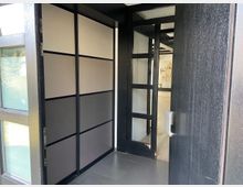 Ansicht eines modernen Eingangsbereichs mit dunklem Holzrahmen, einer Glastür mit mehreren rechteckigen Fenstereinsätzen und einer Wand mit vertikal geteilten Paneelen in neutralen Farbtönen. Im Hintergrund ist ein Flur mit hellem Boden und ein gemaltes Porträt an der Wand zu sehen.