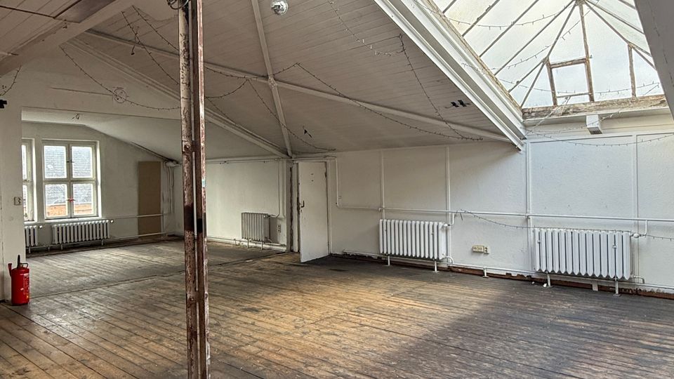Großer, leerer Dachgeschossraum mit breitem Oberlicht aus Glas im schrägen Dach, weißen Wänden und sichtbaren Holzbalken. Alter Holzdielenboden, mehrere Heizkörper entlang der Wände und eine einzelne Stützsäule in der Raummitte.