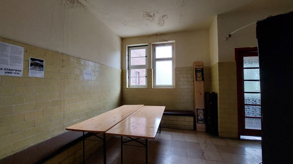 Ein schlichter Raum mit gelblichen Wandfliesen, zwei Fenstern und einer Glastür. Im Vordergrund steht ein Holztisch auf Metallgestell, an der Wand befindet sich eine Bank, und ein zusammenklappbarer Tisch oder Gegenstand lehnt an der Ecke.