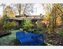 Ein bewachsener Garten mit einer Vielzahl von Pflanzen und Bäumen, im Hintergrund ein flaches Haus mit Wellblechdach und großen Fenstern. Im Vordergrund stehen drei blaue Metallstühle auf einer Wiese mit vereinzeltem Laub.