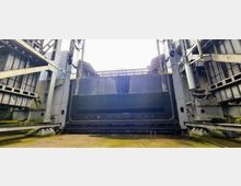 Ansicht eines massiven Metalltores in einem Industrie- oder Schleusenbauwerk, flankiert von stählernen Trägern und Strukturen. Der Boden ist mit Betonplatten bedeckt, während das Tor oben Licht durchscheinen lässt, was auf eine offene Deckenstruktur hinweist.