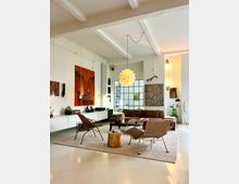 Open-plan modern living room with white walls and exposed ceiling beams, featuring a large grid-paned window and contemporary pendant lighting. The space includes wall art, low built-in cabinets, a brown sofa with lounge chairs on a large rug, and minimal decor on a polished light floor.