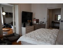 Ein Schlafzimmer mit einem großen weißen Bett im Vordergrund, einem Flachbildfernseher an der Wand und einer Kommode mit Lautsprechern und Deko-Objekten. Im Hintergrund sind ein angrenzendes Wohnzimmer mit Holzboden und Möbeln sowie eine weitere Tür zu einem Nebenzimmer erkennbar.