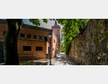 Pflasterweg zwischen einer Backsteinmauer auf der rechten Seite und einem roten Backsteingebäude auf der linken Seite mit einem schwarzen Laternenpfahl; im Hintergrund ein historischer Rundturm und Bäume.
