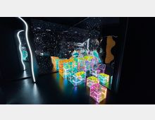 Ein dunkler Raum mit reflektierenden Wänden, die Sterne simulieren, und leuchtenden, farbigen Kuben in verschiedenen Größen, die auf dem Boden angeordnet sind. Die Wände zeigen verzerrte Spiegelungen der Umgebung und verstärken den futuristischen Eindruck.