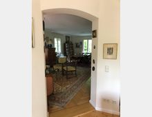 Blick durch einen offenen Türbogen in ein Wohnzimmer mit klassischer Einrichtung, darunter ein großer orientalischer Teppich, ein runder Esstisch mit Stühlen und ein Holzschrank vor einer Wand mit Fenstern. Das Zimmer ist hell durch einfallendes Tageslicht.