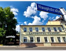 Gelbes Gebäude mit zwei Stockwerken, das die Aufschrift „Landhotel Classic“ trägt, mit weißen Fensterrahmen und Blumenkästen. Im Vordergrund ist ein Straßenschild mit der Aufschrift „Hauptstraße“ sichtbar, während im Hintergrund ein großer Baum und blauer Himmel mit weißen Wolken zu sehen sind.