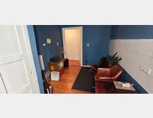 Kleiner Wohnraum mit blauen Wänden, einer weißen Fliesenwand auf der rechten Seite, Holzboden und einem Teppich. Im Raum befinden sich ein brauner Ledersessel, ein kleiner Holztisch mit Laptop, ein Regal mit Plattenspieler sowie eine geöffnete Tür, die in einen hellen Flur führt.