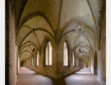 Gewölbter Klostergang mit Spitzbögen und gotischer Architektur, flankiert von hohen, schmalen Fenstern mit Blick auf den Innenhof. Der Gang hat eine gedeckte Ziegeldecke und einen Boden aus roten Fliesen.