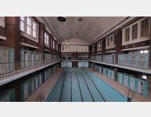 Historisches Hallenbad mit leerem Schwimmbecken, umgeben von zweistöckigen Umkleideräumen in hellblauer Farbe und Wänden aus rotem Backstein; darüber eine hohe Decke mit großen Fenstern und hängenden Lampen.