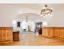 Heller Innenraum mit weißer Tonnenwölbung und großen Rundbögen, Holzdielenboden und umlaufender Holzvertäfelung. Mehrere Kronleuchter und antike Holzschränke stehen entlang der Wände, im Hintergrund sind ein Fenster und ein Durchgang zu weiteren Räumen sichtbar.