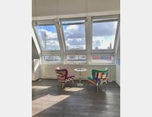 Bright loft seating area with slanted skylight windows overlooking a city skyline, including a tall TV tower and distant domes. Two colorful upholstered chairs face a small round pedestal table on a dark floor beneath hanging pendant lights.