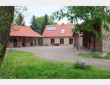 Eine ländliche Hofanlage mit drei Backsteingebäuden, roten Ziegeldächern und Solarzellen auf einem der Dächer. Der Innenhof ist mit Kies bedeckt und von Wiese und Bäumen umgeben.
