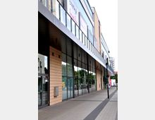 Außenansicht eines modernen Einkaufszentrums mit großen Glasfassaden, beige verkleideter Fassade und einer überdachten Eingangszone. Im Hintergrund sind Schilder von Geschäften sowie Bäume und eine Straße erkennbar.