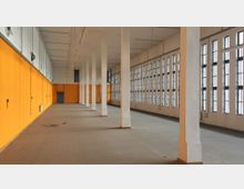 Industriehalle mit gelb-orange gestrichenen Wänden, einer langen Reihe weißer Säulen und hohen Fenstern mit schwarzen Rahmen auf der rechten Seite. Der Boden ist grau und die Decke schlicht mit hängenden Leuchtstofflampen.