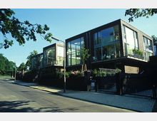 Moderne Wohnhäuser mit großen Glasfronten und dunkel verklinkerten Fassaden, gelegen an einer ruhigen Straße mit Gehweg und üppiger Begrünung im Hintergrund.