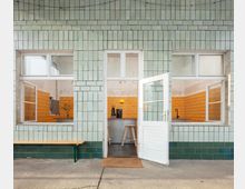 Fassade mit hellgrünen Fliesen und einer offenen weißen Tür, die in einen Innenraum mit orangefarbenen Fliesen und moderner Einrichtung führt. Zwei Fenster flankieren die Tür, während außen eine schlichte Holzbank vor der Wand steht.