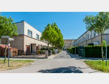 Straßenansicht eines Wohngebiets mit modernen Reihenhäusern aus hellen Ziegeln, flankiert von gepflegten Bäumen und Gehwegen. Im Hintergrund steht ein gelbes, mehrstöckiges Gebäude; Straßenschilder weisen die Namen "Nobelstraße" und "Korczakstraße" aus.