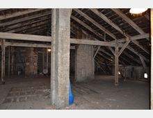 Alter Dachboden mit sichtbaren Holzbalken und einem teilweise beschädigten Holzboden. Die Umgebung wirkt verlassen, mit freistehenden Betonstützen und einem blauen Eimer als prominentem Element im Vordergrund.