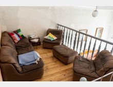 Gemütlicher Wohnbereich auf einer Galerie mit braunen Polstermöbeln, Holzboden und einem kleinen Beistelltisch mit Büchern. Ein Geländer mit gedrechselten Stäben trennt den Bereich von einem offenen Raum darunter, der teilweise sichtbar ist.